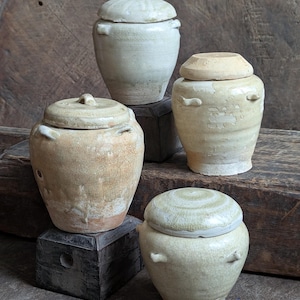 May include: Four antique ceramic jars with lids. The jars are in varying shades of cream and beige, with visible signs of age and wear. Each jar has a rounded body and a lid that fits snugly on top. The jars are arranged on a rustic wooden surface.