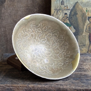 May include: A large, round, silver bowl with a decorative floral pattern. The bowl has a gold rim and is sitting on a wooden surface.