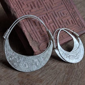 Puede incluir: Un par de pendientes de aro de plata con intrincados diseños florales. Los pendientes están hechos de un metal grueso y pesado y tienen una forma ligeramente curvada.