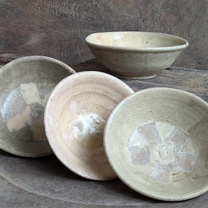 May include: A collection of antique ceramic bowls in varying sizes and designs. The bowls have a neutral color palette with visible wear and tear, suggesting age. Some bowls feature faded, geometric patterns.