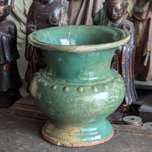 May include: A green ceramic vase with a rounded body and a flared rim. The vase has a speckled glaze and a slightly chipped rim.