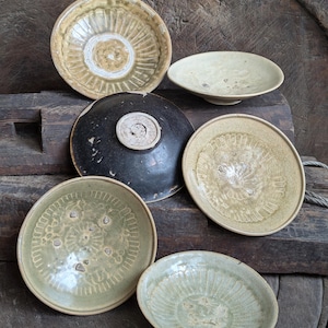 Puede incluir: Una colección de cuencos de cerámica antiguos de varios tamaños y colores. Los cuencos varían del verde claro al beige y negro, con patrones y texturas decorativas. Algunos cuencos tienen un centro elevado, mientras que otros tienen un diseño liso y poco profundo. Los cuencos están dispuestos sobre una superficie de madera.
