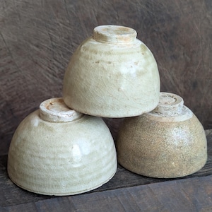 Puede incluir: Tres cuencos de cerámica con forma redondeada y base con pie. Los cuencos presentan un acabado de esmalte beige claro agrietado con sutiles variaciones de tono y textura en sus superficies, mostrando un perfil exterior suave y curvado.