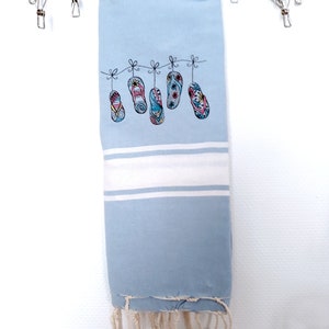 May include: A light blue beach towel with a white stripe and fringe. The towel is decorated with an embroidered design of colorful flip-flops hanging on a line. The towel is hanging from a metal drying rack.