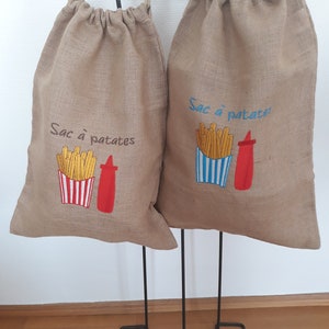 May include: Two beige burlap drawstring bags with embroidered designs. One bag has "Sac à patates" and a fries and ketchup design. The other bag has the same text and design. The bags are hanging on a black metal stand.