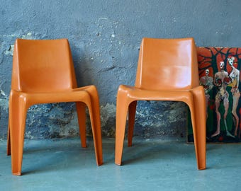 May include: Two orange plastic chairs with a unique, modern design. The chairs are placed in front of a gray wall and a painting with figures.