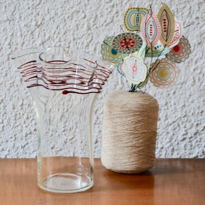May include: A clear glass vase with a ruffled top and red and white striped design. The vase is filled with a brown yarn spool and colourful paper flowers.