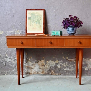 Peut inclure: Une console en bois de style vintage avec trois tiroirs et des pieds effilés. La table est en bois brun clair. Une image encadrée, une petite boîte et un vase avec des fleurs violettes sont posés dessus. La table mesure 120 cm de long.