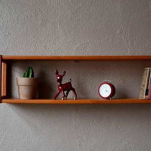 May include: A wooden shelf displaying a cactus in a pot, a red deer ornament, a red alarm clock, and two books. The shelf is mounted on a grey textured wall. The items are arranged to create a decorative display for a home.