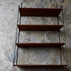 May include: A wall-mounted shelving unit with four wooden shelves and black metal supports. The shelves are a dark brown colour, and the metal frame has a minimalist design. The unit is against a textured, mottled grey wall.