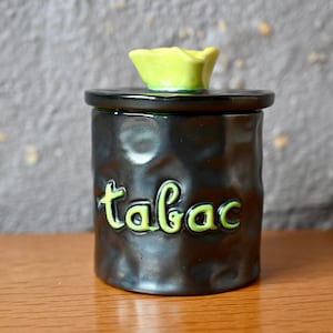 May include: A black ceramic tobacco jar with a green lid and the word "tabac" written in green on the front.