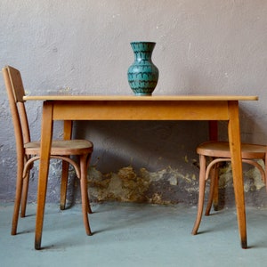 Peut inclure: Une table en bois avec deux chaises et un vase bleu turquoise à motifs noir et blanc. La table et les chaises sont en bois clair et ont un design simple.