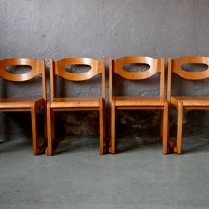 May include: Four wooden chairs with a unique oval cutout in the backrests. The chairs are arranged in a row and are made of light brown wood.