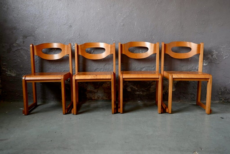 May include: Four wooden chairs with a unique oval cutout in the backrests. The chairs are arranged in a row and are made of light brown wood.