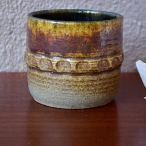 以下が含まれることがあります： テクスチャ加工された縁と中央に隆起したドットのバンドが付いた、茶色と黄色のセラミックマグカップです。