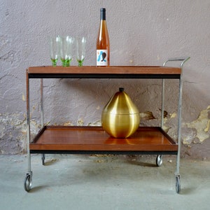May include: A vintage bar cart with a wooden top and chrome frame. The cart has two shelves and four wheels. A bottle of wine, four glasses, and a gold ice bucket are on the top shelf.