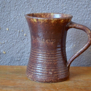 Peut inclure: Une tasse en céramique marron faite à la main avec une anse. La tasse a une surface texturée avec des lignes horizontales et un dessus légèrement évasé. L'intérieur et le bord ont une teinte jaune-brun. La tasse est probablement utilisée pour boire des boissons.