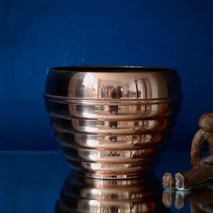 Peut inclure: Un bol en cuivre poli avec une série de rainures horizontales entourant l'extérieur. Le bol a une surface réfléchissante et une forme arrondie. À côté du bol se trouve une petite figurine brune assise avec une posture voûtée et une tête ronde.