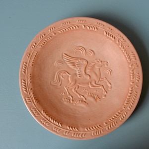 May include: A terracotta plate featuring a raised design of a winged horse, surrounded by a border of stylised leaves.