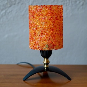 May include: A table lamp featuring a cylindrical shade made of textured, translucent orange and red plastic chips. The lamp has a black base with three curved legs, a brass-colored neck, and a black socket holding the shade in place.