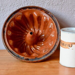 May include: An antique brown bundt cake pan with a central hole and a decorative, ridged design. Beside it is a white tin with a label that reads "remont". The items are on a wooden surface.