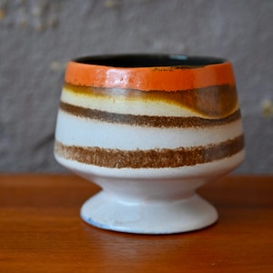 Puede incluir: Un macetero de cerámica blanca con rayas marrones y naranjas. El macetero tiene una base ancha y una parte superior estrecha.