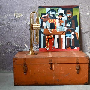 Op de afbeelding: Een vintage houten kist met messing beslag, een gouden trompet en een ingelijst schilderij van vier figuren die muziekinstrumenten bespelen. Het schilderij is in kubistische stijl met felle kleuren en geometrische vormen.