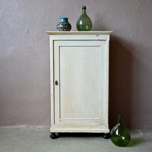 Peut inclure: Un meuble ancien blanc cassé à une porte, surmonté d'une bouteille en verre vert et d'un petit vase à motifs. Une autre bouteille en verre vert est posée au sol. Le meuble a des pieds noirs.