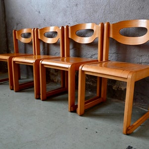 May include: Four wooden chairs with a simple, mid-century modern design. The chairs are made of light brown wood and have a curved back with a cutout. The chairs are arranged in a row.
