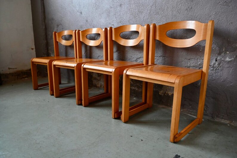 May include: Four wooden chairs with a simple, mid-century modern design. The chairs are made of light brown wood and have a curved back with a cutout. The chairs are arranged in a row.
