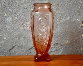 Op de afbeelding: Een perzikroze glazen vaas in Art Deco-stijl. De vaas heeft een uitlopende bovenkant en een gedetailleerd, wervelend ontwerp aan de voorkant. De vaas is ongeveer 25 cm hoog.