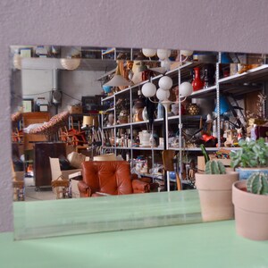 Peut inclure: Un miroir reflétant une pièce encombrée avec des étagères remplies de divers objets, y compris des lampes, des vases et d'autres objets de décoration. Il y a aussi plusieurs plantes en pot au premier plan.