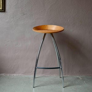 May include: A modern bar stool with a light wood seat and chrome legs. The seat is approximately 14 inches in diameter. The legs are sleek and curved, with a black support bar. The stool is set against a neutral-colored wall.