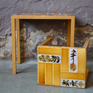 Peut inclure: Deux petites tables en bois. L'une est une simple table carrée avec une finition en bois clair. L'autre est une petite table carrée avec un motif de carreaux jaunes avec des motifs floraux et de feuilles. Les tables sont posées contre un mur gris texturé.