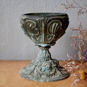 Könnte beinhalten: Ein antiker, verzierter Metall-Pflanztopf mit einem detaillierten, geprägten Design. Der Topf ist dunkelgrün und hat einen breiten, runden Boden.