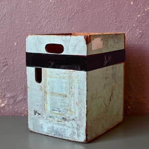 Puede incluir: Una caja de madera vintage con un acabado de pintura azul claro. La caja tiene una banda de cinta negra alrededor y una etiqueta escrita a mano que dice "8 oz".