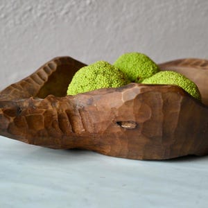 Könnte beinhalten: Eine dunkelbraune, handgeschnitzte Holzschale mit rauer, strukturierter Außenseite und einem länglichen, spitz zulaufenden Ende. In der Schale liegen drei hellgrüne, texturierte, kugelförmige Objekte, die an Früchte des Milchorangenbaums erinnern.