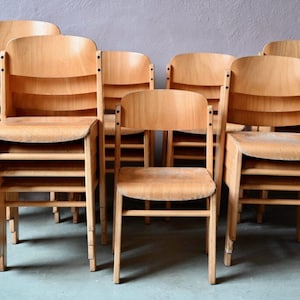 Può includere: Una pila di sedie in legno con una finitura marrone chiaro. Le sedie sono disposte a zig-zag, con la sedia anteriore leggermente spostata rispetto alle altre. Le sedie sono realizzate in legno massello e hanno un design semplice e classico.