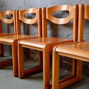 May include: Four wooden chairs with a simple design, featuring a curved back and a cutout in the back. The chairs are arranged in a row and are made of light brown wood.