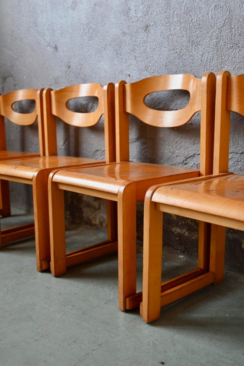 May include: Four wooden chairs with a simple design, featuring a curved back and a cutout in the back. The chairs are arranged in a row and are made of light brown wood.