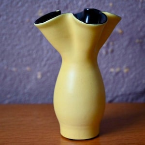 May include: A yellow ceramic vase with a distinctive, frilled rim and a black interior. The vase has a slender, hourglass shape and sits on a wooden surface. The background is a textured purple wall.