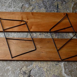 May include: Two wooden shelves with a natural finish and black metal geometric supports. The shelves are rectangular and have a warm, reddish-brown hue. The metal supports are angular, creating a modern aesthetic.
