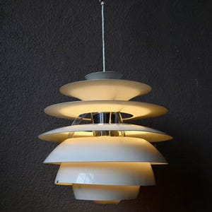 Peut inclure: Un luminaire suspendu blanc à plusieurs niveaux avec un centre chromé. La lumière est allumée et éclaire la pièce.