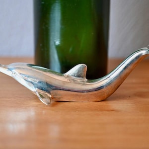 May include: A silver-toned metal bottle opener shaped like a dolphin. The dolphin is depicted in a horizontal swimming position with its tail curved upward, forming a loop at the end. The surface of the metal is smooth and reflective.