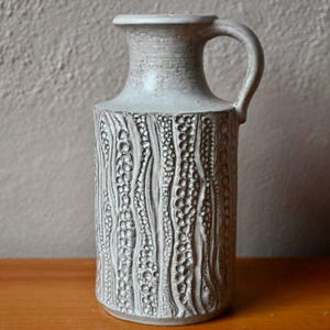 May include: A textured, off-white ceramic vase with a handle. The vase has a cylindrical body with a detailed, raised pattern. The handle curves gracefully from the top to the side. The vase is on a wooden surface.