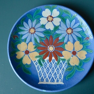 May include: A blue ceramic plate featuring a painted design of a bouquet of flowers in a basket. The flowers are yellow, white, blue, and red.