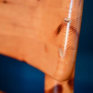May include: Close-up of the back of a wooden chair with a natural wood finish. The chair has a curved back and a smooth, polished surface.
