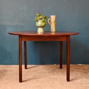 Peut inclure: Une table ovale en bois foncé avec un motif de grain de bois clair. La table a un design simple avec quatre pieds et un dessus lisse. Deux vases en céramique sont sur la table, un vert et un autre avec un motif coloré.