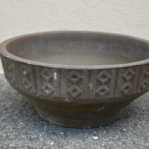 Puede incluir: Una maceta de terracota marrón con un patrón geométrico alrededor del borde. La maceta está sobre una superficie gris.