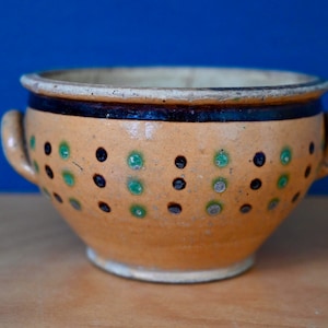 Puede incluir: Un pequeño cuenco de cerámica marrón con dos asas. El cuenco está decorado con un patrón de lunares negros y verdes.